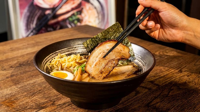 A hand uses chopsticks to pick up a slice of pork from a bowl of ramen with noodles and egg.