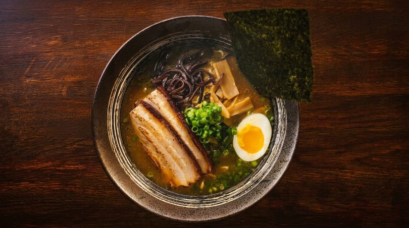 Overhead view of a ramen bowl with pork belly, egg, seaweed, and green onions.