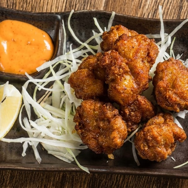 Crispy fried chicken pieces with shredded cabbage, a lemon wedge, and orange dipping sauce on a dark plate.