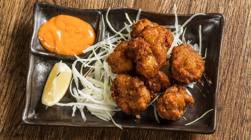 Crispy fried chicken pieces with shredded cabbage, a lemon wedge, and orange dipping sauce on a dark plate.