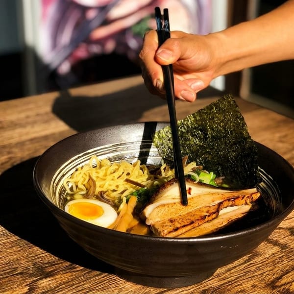 A hand uses chopsticks to pick up ramen noodles from a black bowl on a wooden table.