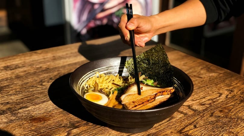A hand uses chopsticks to pick up ramen noodles from a black bowl on a wooden table.