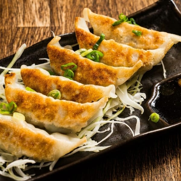 Golden-brown gyoza dumplings with shredded cabbage and dipping sauce on a dark rectangular plate.