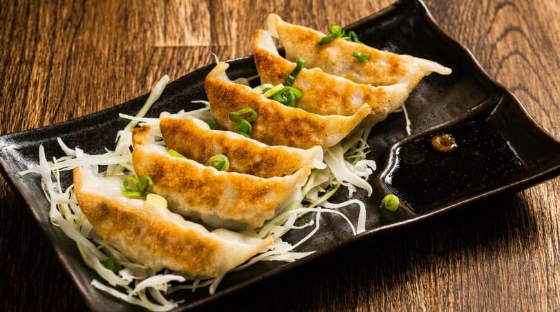 Golden-brown gyoza dumplings with shredded cabbage and soy sauce on a dark rectangular plate.