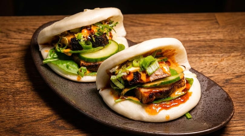 Two bao buns filled with meat, cucumber, greens, and sauces on a dark plate.